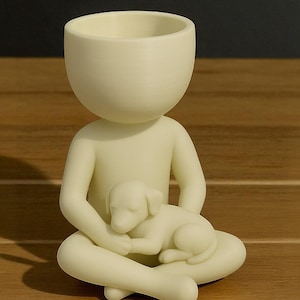 May include: A cream-colored ceramic planter shaped like a person seated with legs crossed, cradling a sleeping puppy. The planter has a large, open head for planting. The figurine is on a wooden surface.