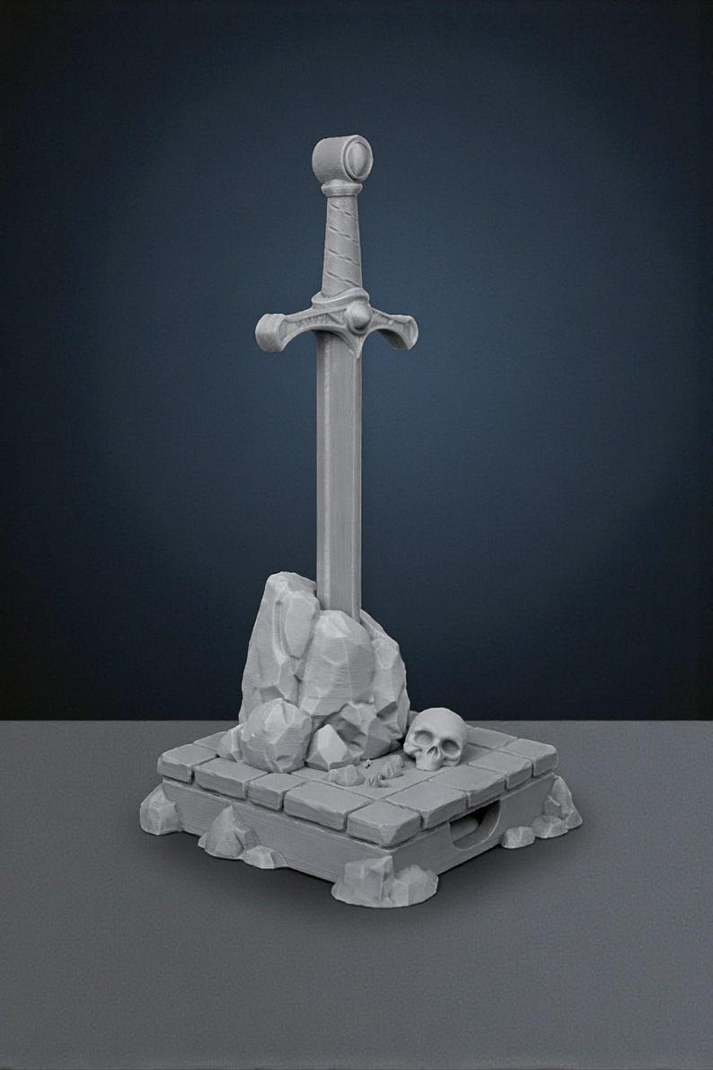 May include: A gray sword in stone sculpture. The sword is upright, with a detailed hilt and crossguard. The stone base features a skull and rock formations, creating a fantasy theme. The sculpture is made of a solid, gray material.