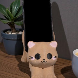 May include: A tan cat-shaped phone holder with pink inner ears and black eyes, holding a black smartphone. The holder sits on a wooden surface, with a small potted plant and a cup in the background.