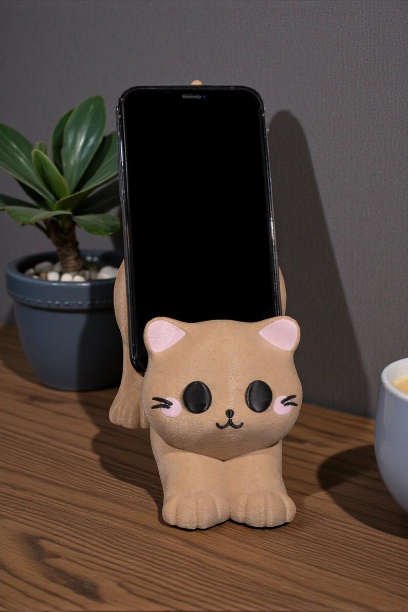 May include: A tan cat-shaped phone holder with pink inner ears and black eyes, holding a black smartphone. The holder sits on a wooden surface, with a small potted plant and a cup in the background.