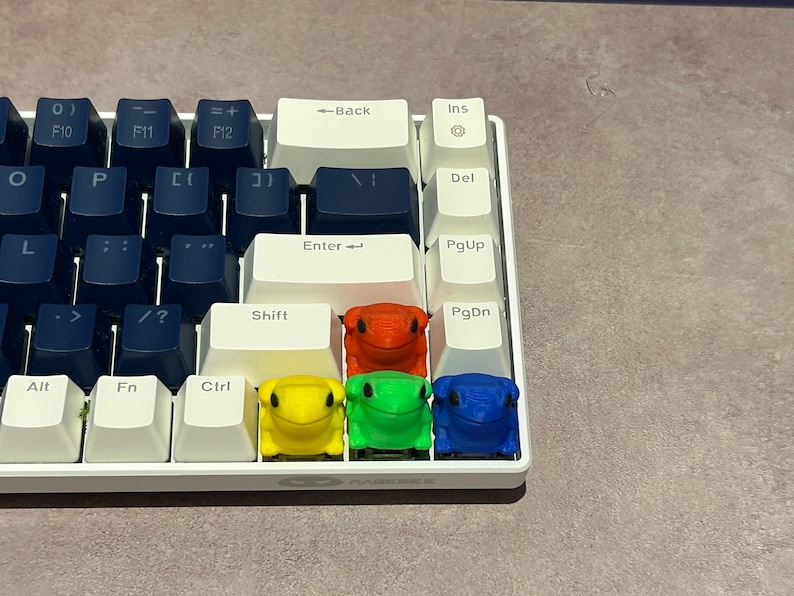 May include: A white keyboard with four frog-shaped keycaps. The keycaps are red, yellow, green, and blue. The keyboard has keys labelled 'Enter', 'Shift', 'Alt', 'Fn', 'Ctrl', 'PgUp', 'PgDn', 'Ins', 'Del', 'Back', 'F10', 'F11', 'F12', '0)', '=', '-', '[', ']', '/', '>', '?', ';', '"', ',', '.', 'L', 'O', 'P'.
