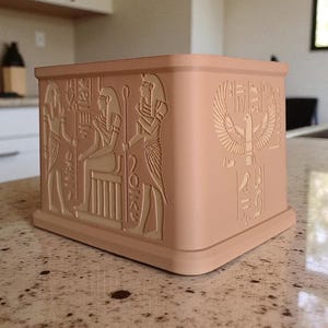 May include: A square, light-brown decorative box with detailed relief carvings of Egyptian figures and hieroglyphs. The box features a base and a slightly curved top edge. The carvings include a winged sun disk and seated figures.