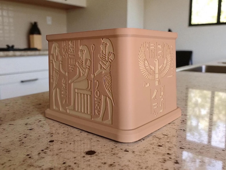 May include: A square, light-brown decorative box with detailed relief carvings of Egyptian figures and hieroglyphs. The box features a base and a slightly curved top edge. The carvings include a winged sun disk and seated figures.