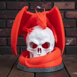 May include: A red dragon figurine perched atop a white skull, with the dragon's wings spread wide. The skull is encircled by a red snake, all set on a gray base. The background features a brick wall and a wooden surface.