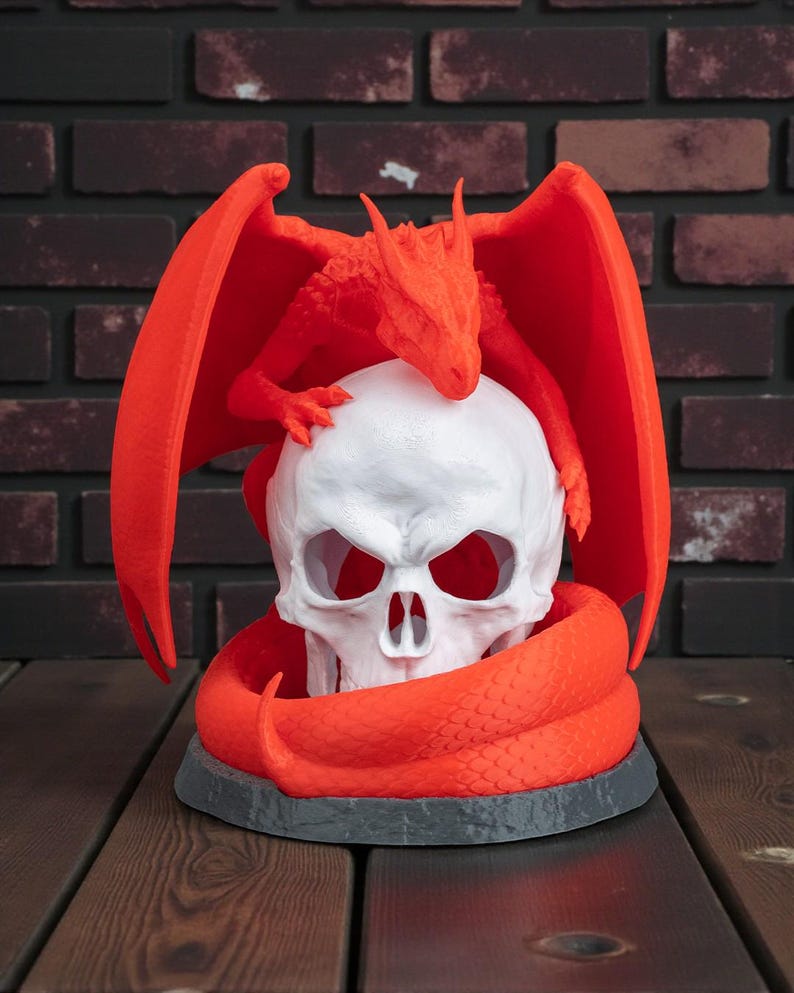 May include: A red dragon figurine perched atop a white skull, with the dragon's wings spread wide. The skull is encircled by a red snake, all set on a gray base. The background features a brick wall and a wooden surface.