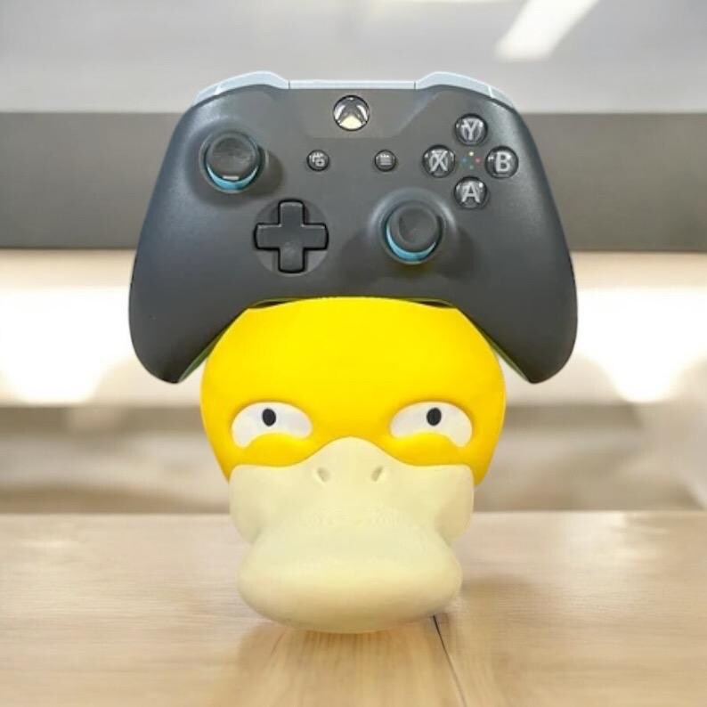 May include: A yellow Psyduck figure with a white beak and eyes, topped with a gray Xbox controller. The controller has blue accents around the joysticks and buttons.