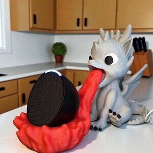 May include: A whimsical dragon-shaped speaker holder in gray and red. The dragon's tongue forms a cradle for a black speaker. The holder is on a white surface, with a kitchen background.