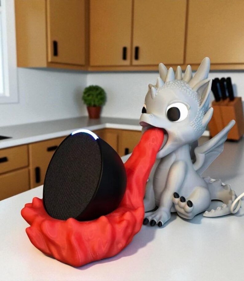May include: A whimsical dragon-shaped speaker holder in gray and red. The dragon's tongue forms a cradle for a black speaker. The holder is on a white surface, with a kitchen background.