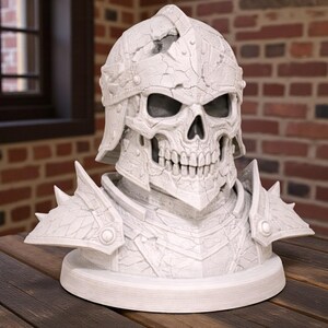 May include: A white, detailed bust of a skull wearing a helmet and armor. The skull has visible teeth and eye sockets. The helmet and armor have a textured, cracked appearance. The bust is on a round base, and the background is a brick wall.