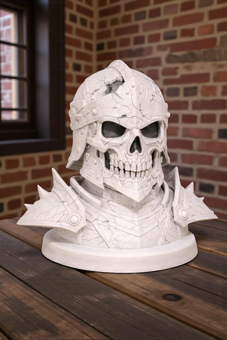 May include: A white, detailed bust of a skull wearing a helmet and armor. The skull has visible teeth and eye sockets. The helmet and armor have a textured, cracked appearance. The bust is on a round base, and the background is a brick wall.