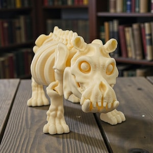 May include: A cream-colored, 3D-printed sculpture of a skeletal creature, possibly a dinosaur or mythical beast. The sculpture has detailed rib and spine structures, a skull-like head with large eye sockets, and four clawed feet. It is posed on a wooden surface.