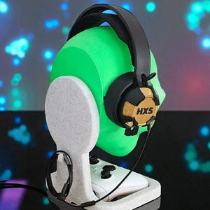 May include: A white headset stand with a green head-shaped form holding a black and gold headset. The stand also holds a white game controller. The headset has the text "HX5" on the ear cup.
