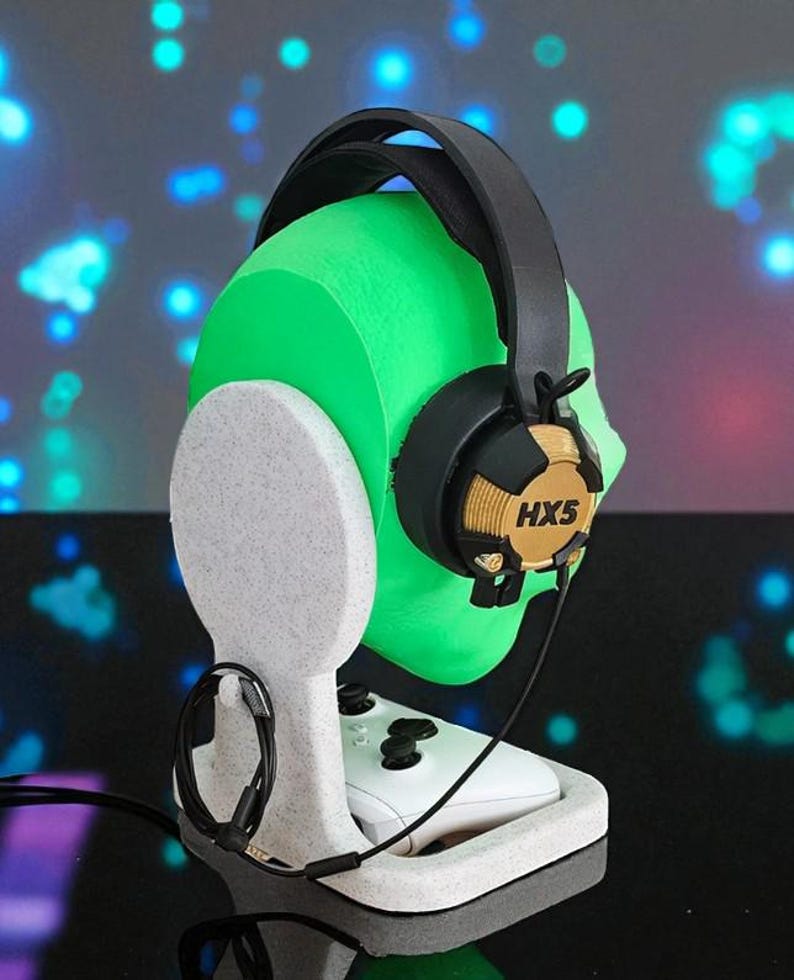 May include: A white headset stand with a green head-shaped form holding a black and gold headset. The stand also holds a white game controller. The headset has the text "HX5" on the ear cup.