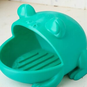 May include: A teal frog-shaped soap dish. The dish has a wide open mouth and a textured interior. The frog has large eyes and a rounded head. The dish is made of a solid material and is designed to hold a bar of soap.