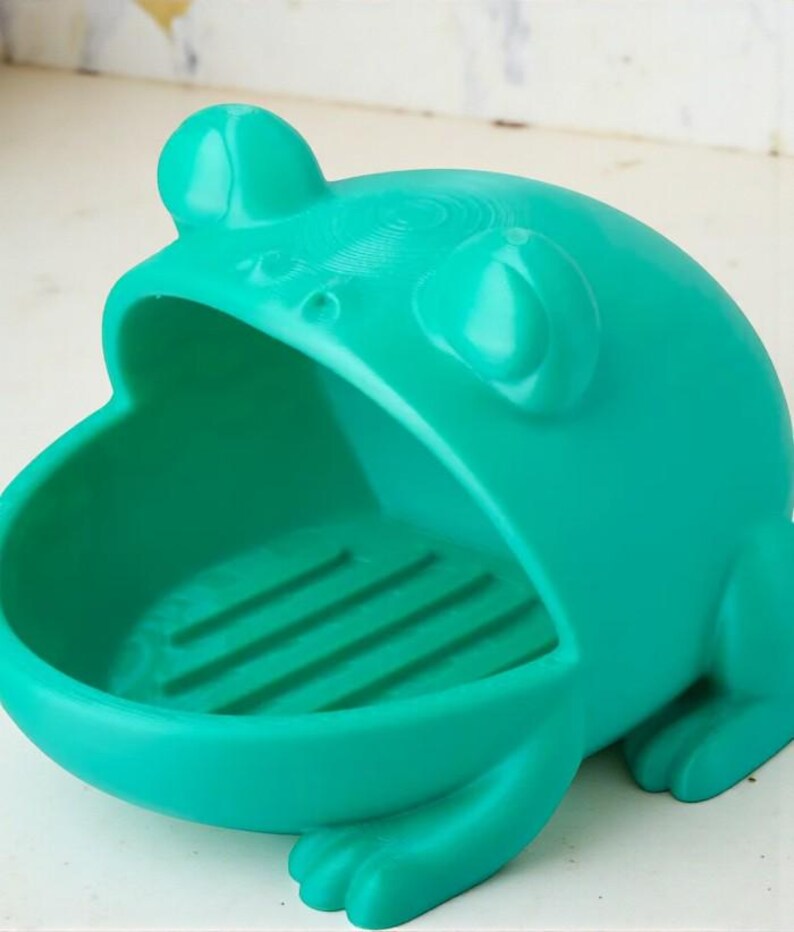 May include: A teal frog-shaped soap dish. The dish has a wide open mouth and a textured interior. The frog has large eyes and a rounded head. The dish is made of a solid material and is designed to hold a bar of soap.