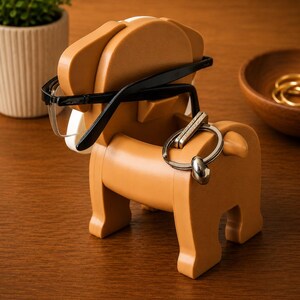May include: A tan-colored dog-shaped organizer holding black-framed eyeglasses and a silver keychain. The organizer has a white accent and is made of a solid material. The dog-shaped organizer is on a wooden surface.