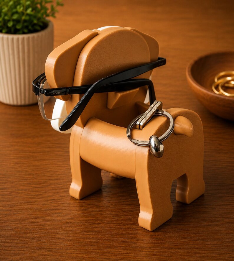 May include: A tan-colored dog-shaped organizer holding black-framed eyeglasses and a silver keychain. The organizer has a white accent and is made of a solid material. The dog-shaped organizer is on a wooden surface.