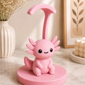 May include: A pink axolotl figurine with a smiling face, sitting on a round base. The figurine has detailed gills and a tail, and is connected to a curved support. The overall design is a cute and whimsical decorative item.