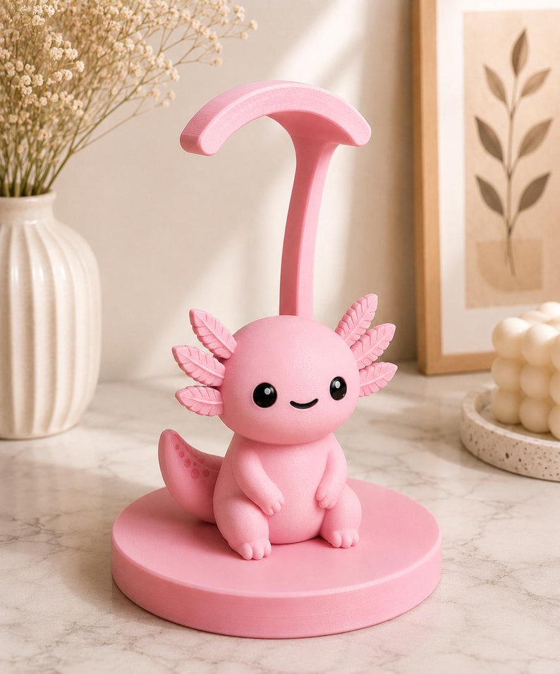 May include: A pink axolotl figurine with a smiling face, sitting on a round base. The figurine has detailed gills and a tail, and is connected to a curved support. The overall design is a cute and whimsical decorative item.