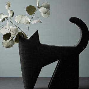 May include: A black cat-shaped vase with a textured surface. The vase holds a sprig of eucalyptus with round, pale green leaves. The cat's tail curves upwards, and its body has geometric cutouts. The vase is on a dark surface.