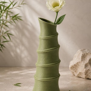 Puede incluir: Un jarr&oacute;n verde con forma de bamb&uacute; que sostiene una flor blanca con un tallo verde. El jarr&oacute;n tiene una superficie texturizada y un dise&ntilde;o curvo &uacute;nico. Un art&iacute;culo decorativo para el hogar.