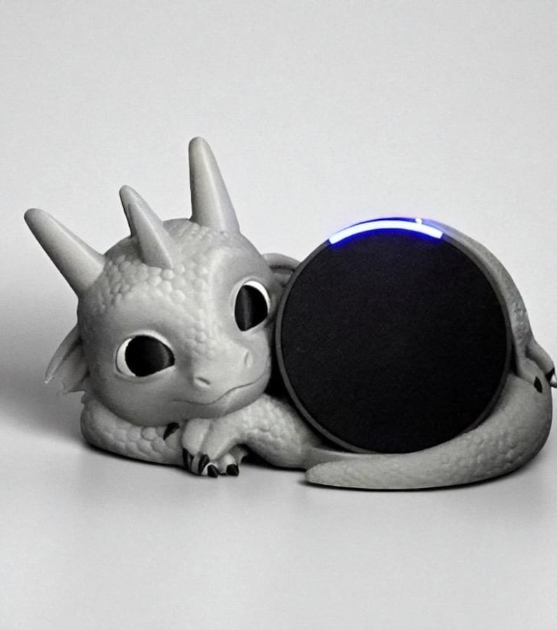 May include: A gray dragon-shaped speaker holder with a round black speaker. The dragon has large eyes, horns, and a textured body. The speaker has a blue light on top. The overall design is whimsical and decorative.