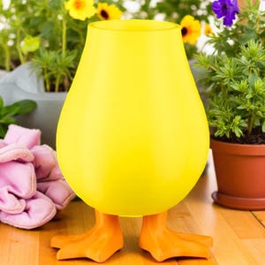 Puede incluir: Un jarr&oacute;n de vidrio amarillo caprichoso con forma de pato, con patas naranjas. El jarr&oacute;n tiene un cuerpo redondeado y una parte superior acampanada. El fondo incluye flores en macetas y una superficie de madera.