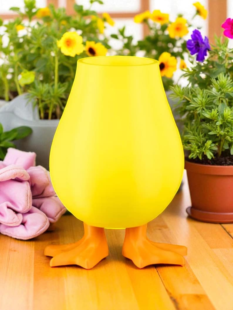 Puede incluir: Un jarr&oacute;n de vidrio amarillo caprichoso con forma de pato, con patas naranjas. El jarr&oacute;n tiene un cuerpo redondeado y una parte superior acampanada. El fondo incluye flores en macetas y una superficie de madera.