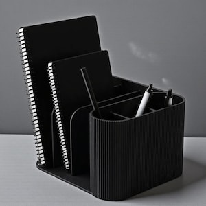 May include: A black desk organizer with multiple compartments. It holds two spiral-bound notebooks, a notepad, and pens. The organizer has a ribbed texture and a sleek, modern design, perfect for office or home use.
