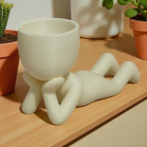 以下が含まれることがあります： クリーム色のセラミックプランター。横たわっている人の形をしており、丸い頭と頭を支える腕があります。プランターには植物用の円形の開口部があります。脚は伸びています。プランターは木製の表面にあります。