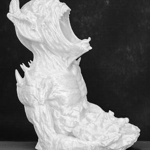 May include: A white sculpture of a creature with a wide open mouth, horns, and sharp teeth. The figure is seated with its arms bent and its body in a hunched position. The sculpture has a rough, textured surface.