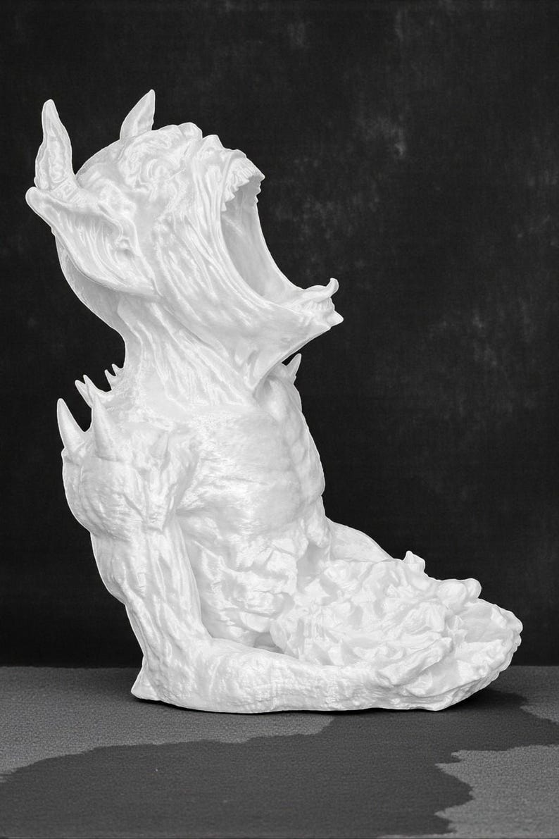 May include: A white sculpture of a creature with a wide open mouth, horns, and sharp teeth. The figure is seated with its arms bent and its body in a hunched position. The sculpture has a rough, textured surface.