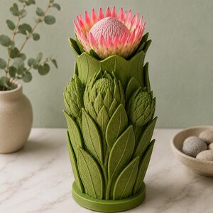 Puede incluir: Un jarr&oacute;n decorativo verde con una flor de protea. El jarr&oacute;n tiene forma de flor estilizada con dise&ntilde;os detallados de hojas y capullos. La flor de protea tiene p&eacute;talos rosas y blancos. El jarr&oacute;n est&aacute; sobre una superficie de m&aacute;rmol blanco.
