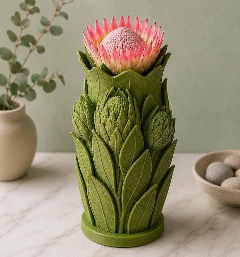 Puede incluir: Un jarr&oacute;n decorativo verde con una flor de protea. El jarr&oacute;n tiene forma de flor estilizada con dise&ntilde;os detallados de hojas y capullos. La flor de protea tiene p&eacute;talos rosas y blancos. El jarr&oacute;n est&aacute; sobre una superficie de m&aacute;rmol blanco.