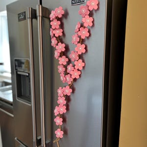 May include: A decorative floral garland with pink blossoms and white centers, attached to a brown stem, is displayed on a stainless steel refrigerator. The garland is a vertical design, adding a touch of spring to the kitchen.