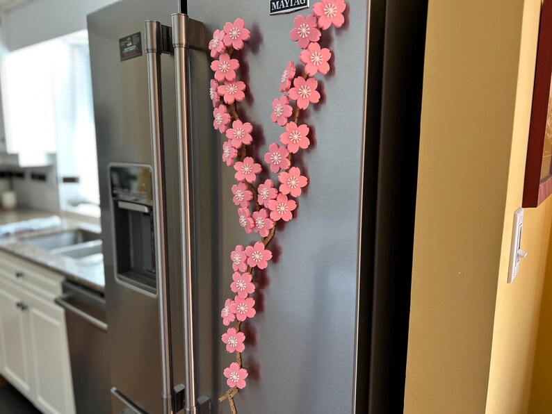 May include: A decorative floral garland with pink blossoms and white centers, attached to a brown stem, is displayed on a stainless steel refrigerator. The garland is a vertical design, adding a touch of spring to the kitchen.