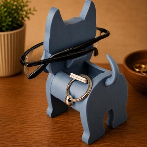 May include: A blue wooden dog-shaped organizer holding black-framed glasses and a silver keyring. The dog has a small compartment for storing small items. The organizer is on a wooden surface.