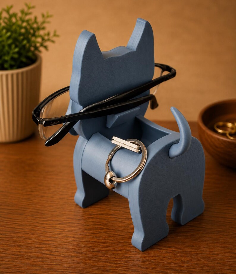 May include: A blue wooden dog-shaped organizer holding black-framed glasses and a silver keyring. The dog has a small compartment for storing small items. The organizer is on a wooden surface.