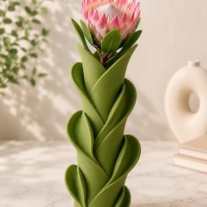 May include: A green, leaf-shaped vase holding a pink and white protea flower. The vase has a unique, layered design. The flower has green leaves and a large, textured bloom. The background includes a white vase and a hint of greenery.