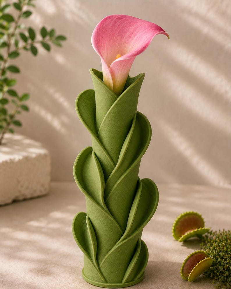 May include: A green, leaf-shaped vase holds a pink calla lily. The vase has a unique, spiraling design. The background includes a small plant and a few Venus flytraps.