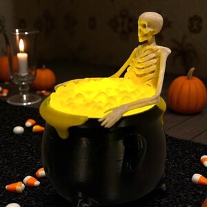 May include: A black cauldron with a yellow glowing liquid and a skeleton figure sitting inside. The cauldron is surrounded by pumpkins, candy corn, and a lit candle in a glass holder, creating a Halloween theme.