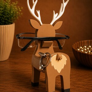 May include: A brown wooden deer-shaped eyeglasses and key holder with white antlers. The holder has a light tan patch on its rear and black feet. Black eyeglasses rest on the deer's nose, and a set of keys hangs from the rear.
