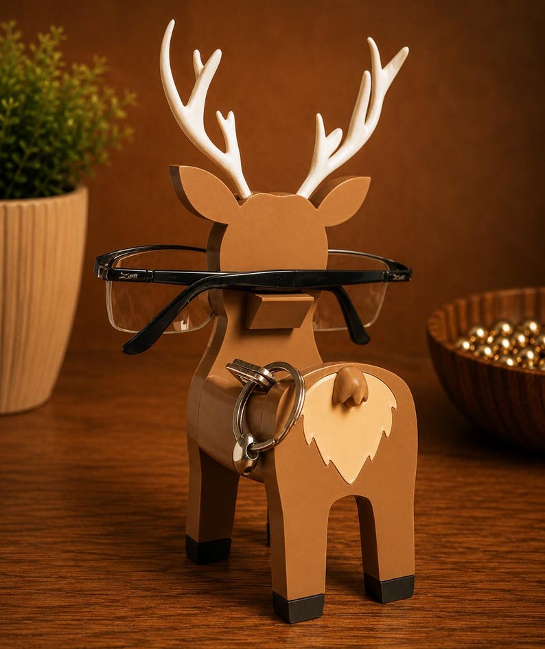 May include: A brown wooden deer-shaped eyeglasses and key holder with white antlers. The holder has a light tan patch on its rear and black feet. Black eyeglasses rest on the deer's nose, and a set of keys hangs from the rear.