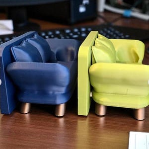 May include: Two miniature armchairs, one navy blue and one lime green, are displayed side-by-side. Each has a rounded seat, back, and armrests, with a small pillow and gold-colored legs. The chairs are set on a wooden surface.