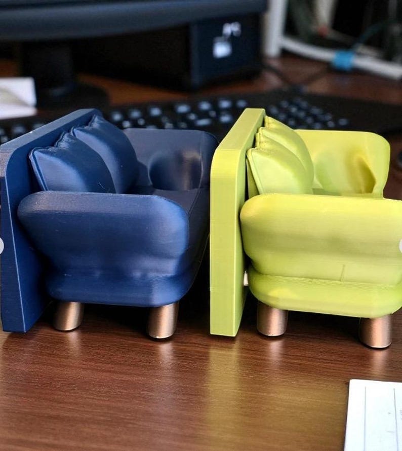 May include: Two miniature armchairs, one navy blue and one lime green, are displayed side-by-side. Each has a rounded seat, back, and armrests, with a small pillow and gold-colored legs. The chairs are set on a wooden surface.