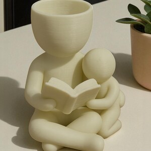 May include: A cream-colored planter shaped like a person reading a book to a smaller figure. The planter has a rounded head and arms, with the book held open in its hands. The smaller figure is nestled beside it. A small plant in a peach pot is in the background.