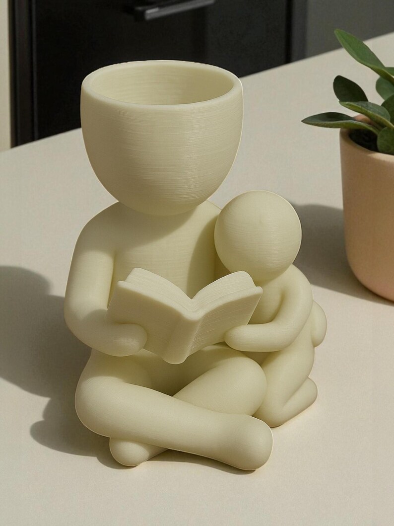 May include: A cream-colored planter shaped like a person reading a book to a smaller figure. The planter has a rounded head and arms, with the book held open in its hands. The smaller figure is nestled beside it. A small plant in a peach pot is in the background.