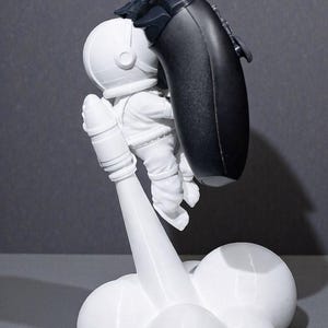 Op de afbeelding: Een witte astronautenfiguur die een zwarte gamecontroller vasthoudt, gemonteerd op een witte raketvormige basis. Het beeldje is gedetailleerd met een helm en ruimtepak. De basis heeft een wolkachtig ontwerp.