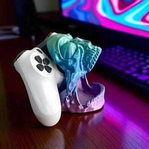 Pu&ograve; includere: Un supporto per controller di gioco a forma di teschio stampato in 3D, nei toni del blu, verde e viola. Un controller di gioco bianco &egrave; nel supporto. Il supporto &egrave; su una superficie di legno.