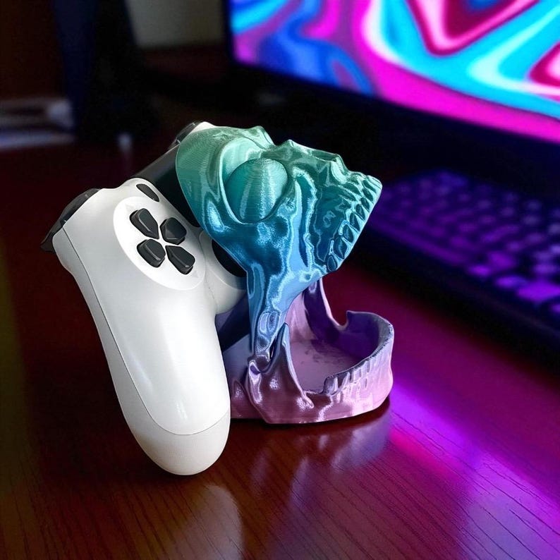 Pu&ograve; includere: Un supporto per controller di gioco a forma di teschio stampato in 3D, nei toni del blu, verde e viola. Un controller di gioco bianco &egrave; nel supporto. Il supporto &egrave; su una superficie di legno.